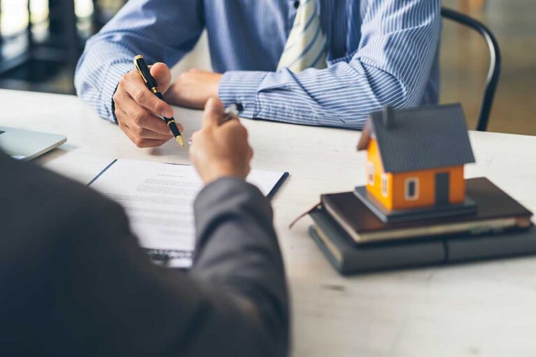 A person’s hand holding new house keys over a wooden table with home documents and a miniature house model.